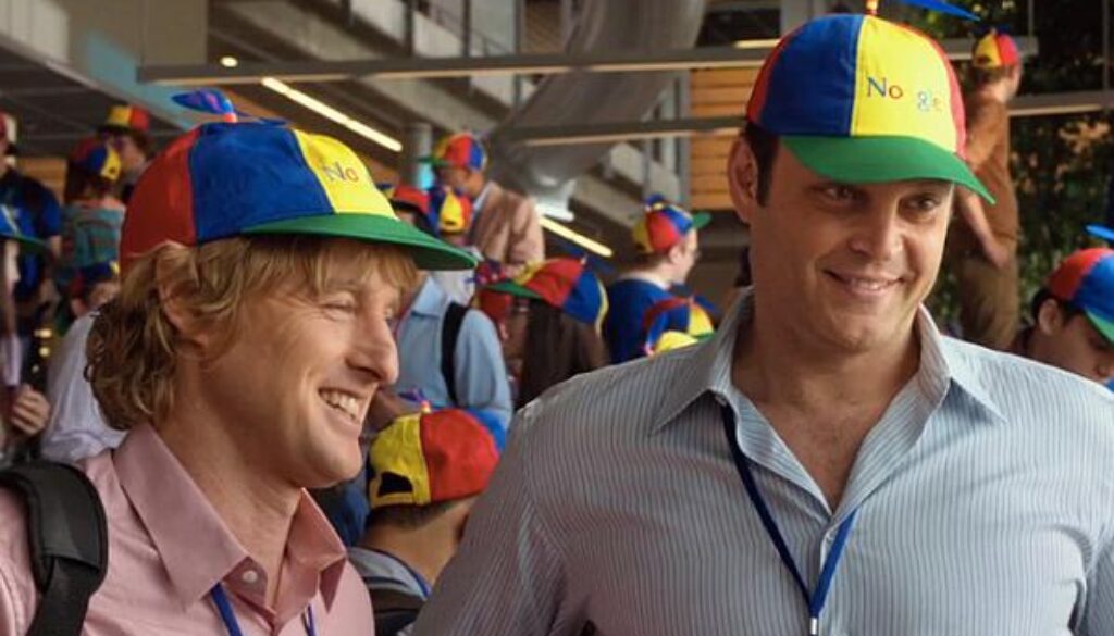 Dos hombres sonrientes con gorras coloridas en un ambiente corporativo concurrido.